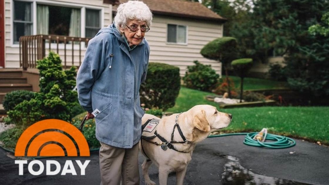 Older adult female with a guide dog in front of her suburban home, wearing blue hooded jacket and glasses, surrounded by neatly trimmed landscaping and beige siding house.