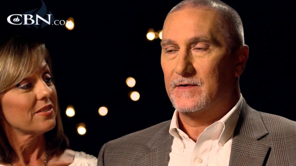 A middle-aged man in a gray blazer and white shirt indoors with blurred lights suggesting an interview setting.