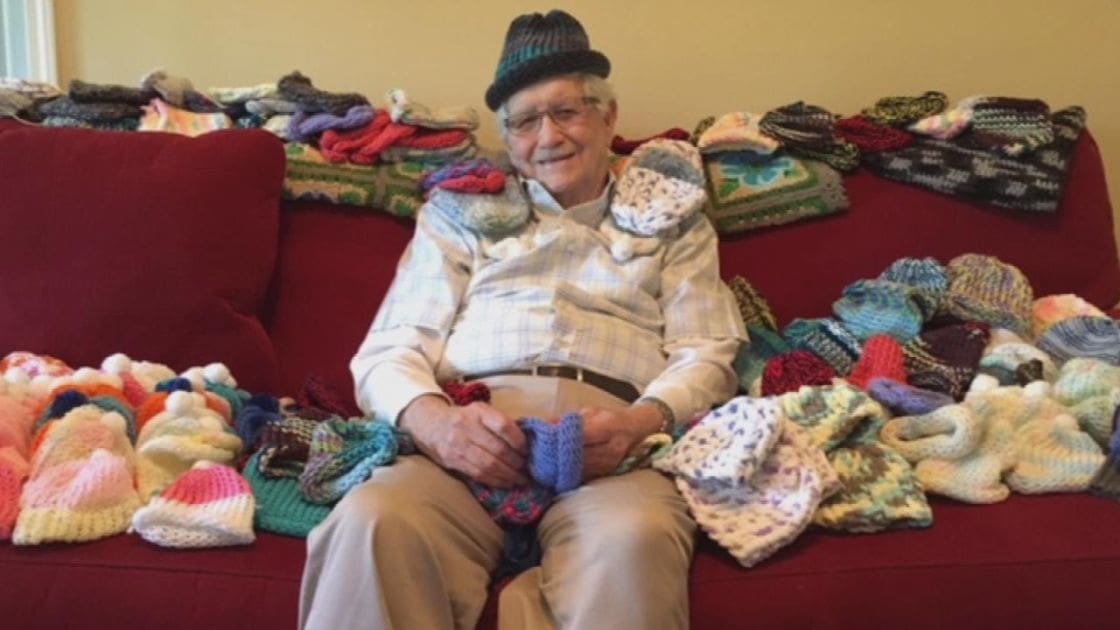 An older adult male sits in a cozy living room, wearing a plaid shirt, beige pants, and a dark green knitted hat with glasses on his face.
