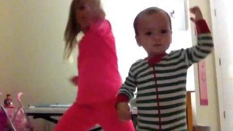 An infant in a green and white striped onesie with red cuffs stands indoors, partially accompanied by another toddler in a bright pink dress or jumpsuit. The setting appears to be a home environment with light-colored walls and toys on the floor.