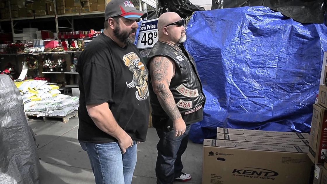 Two adult males in a warehouse setting, one wearing a tiger graphic T-shirt and the other a leather vest over black shirt with '666' patch.