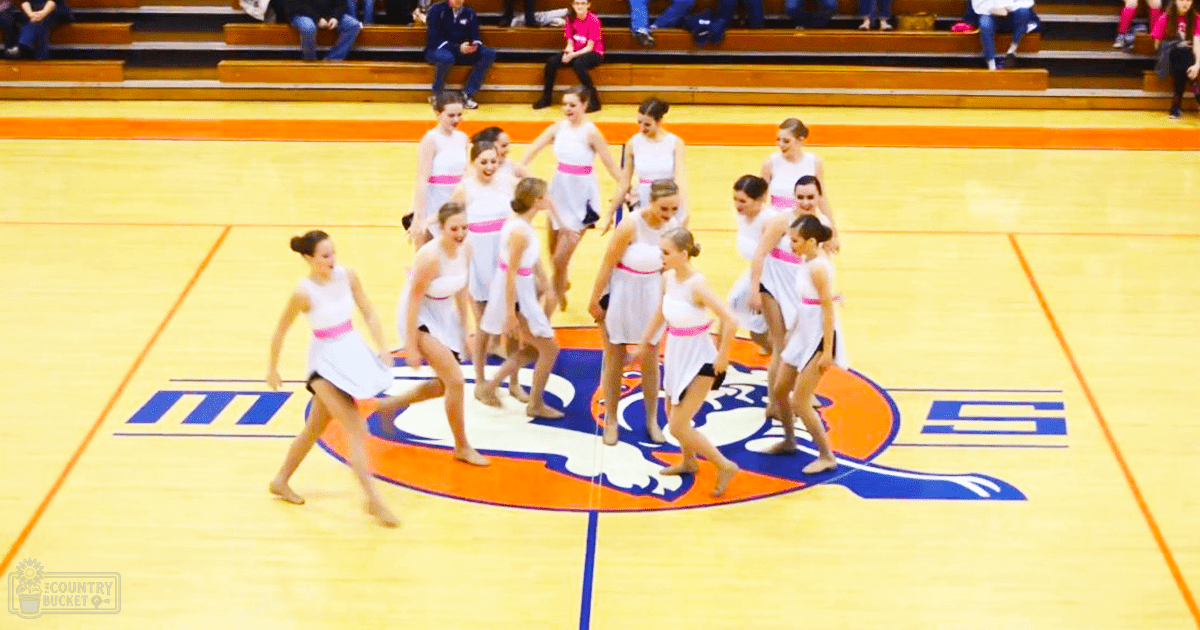 A group of young female dancers in white dresses with pink belts and black undergarments perform barefoot on a basketball court inside a school gymnasium, surrounded by bleachers filled with spectators.