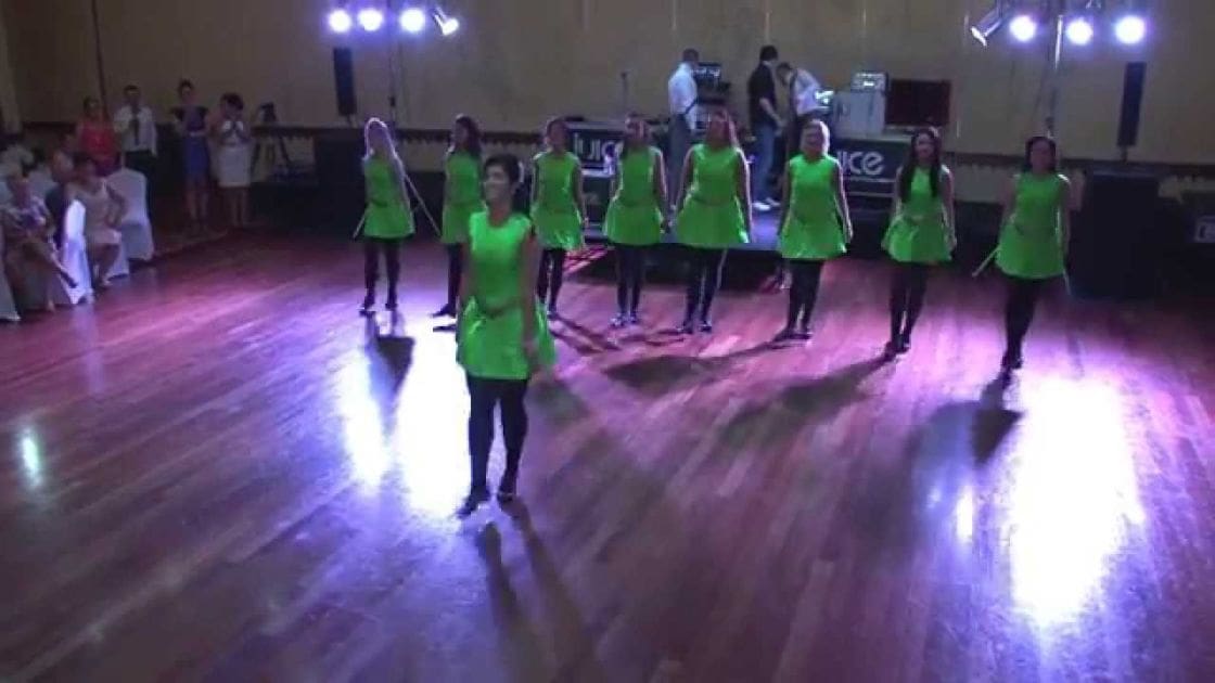 A group of young adult female dancers in bright green tunic dresses perform on a stage within an indoor hall, with spectators dressed formally seated on chairs covered in white tablecloths and sound equipment visible in the background.