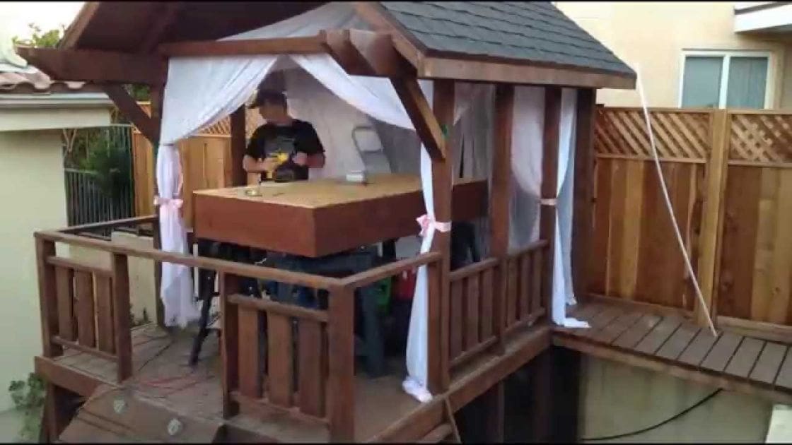 A young adult male playing with a toy gun inside a wooden playhouse in an outdoor residential backyard during daytime, surrounded by greenery.