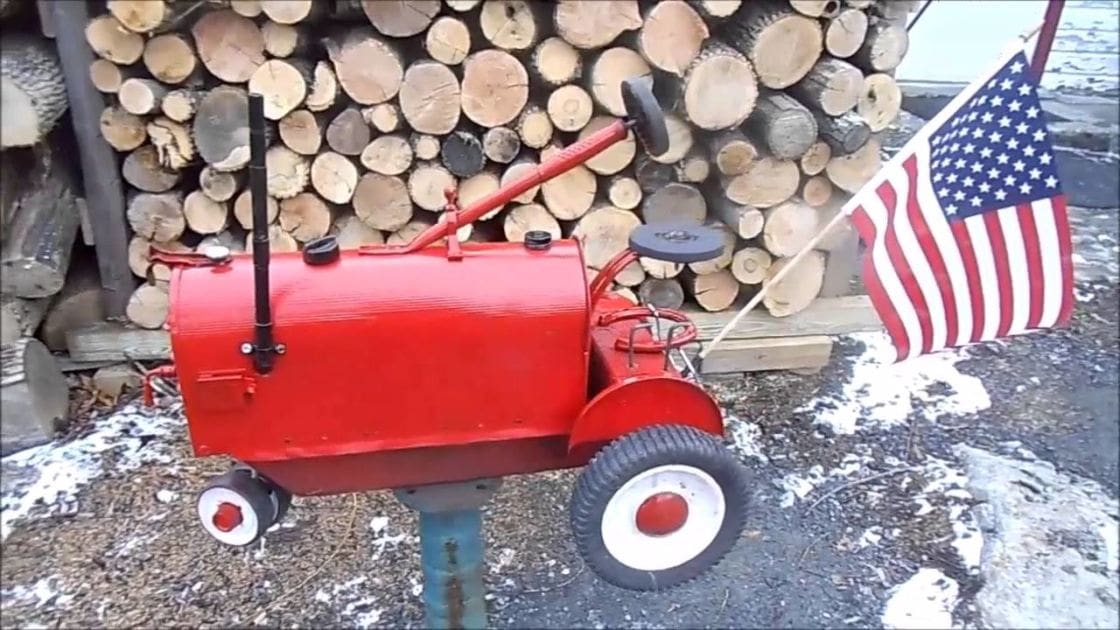 Red toy tractor with white wheels, American flag attached, snow-covered ground in front of logs, overcast light, winter or early spring setting.