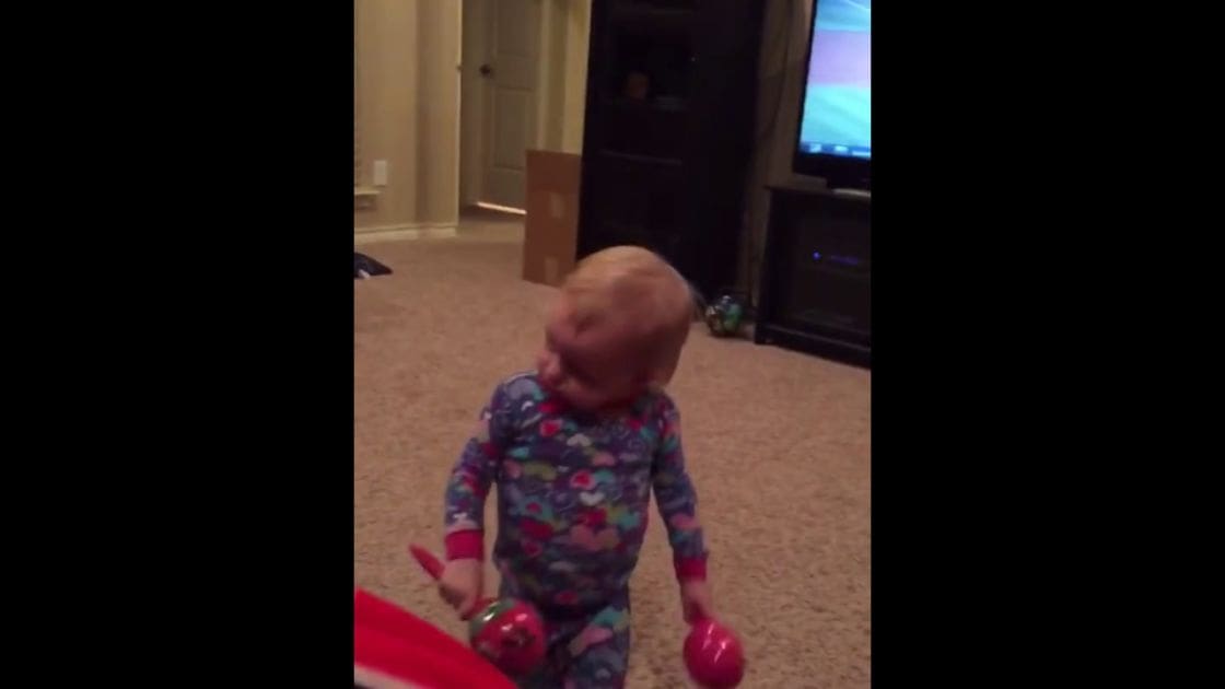 A toddler wearing patterned pajamas in a beige carpeted living room during daytime, holding pink maracas.