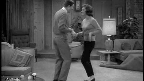 A couple dancing in a cozy mid-20th-century living room with patterned cushions and simple furnishings.