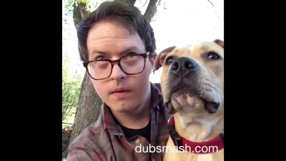 A young adult male wearing a plaid shirt outdoors with an American Pit Bull Terrier mix dog nearby.