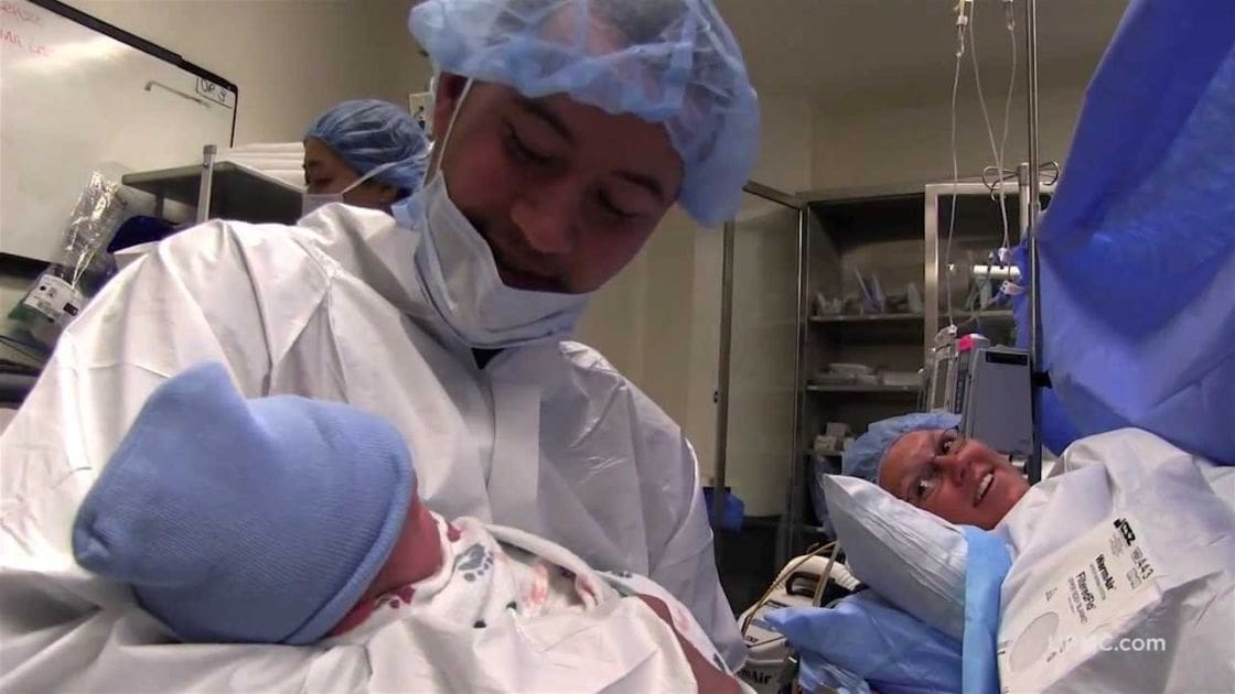 A newborn baby wearing a blue knit hat, held by an adult male in a white surgical gown and light blue hairnet, lying on a hospital bed with other medical personnel present.