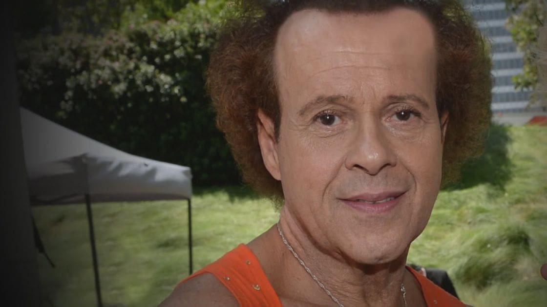 A middle-aged adult male wearing an orange sleeveless top with gold embellishments at the shoulders and neckline, standing in a green outdoor setting with a white tent structure in the background.