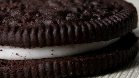 A close-up view of a single Oreo cookie with its signature chocolate wafer and white cream filling. Indoors, bright lighting highlights the texture.