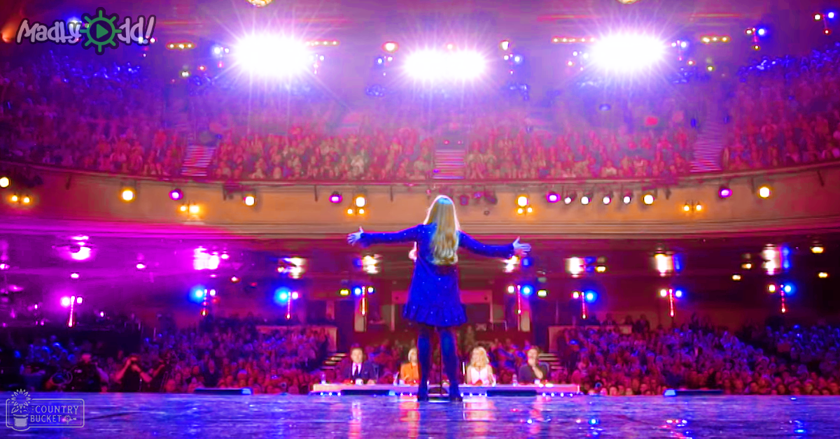 A young adult female performer in a blue textured dress on stage with an audience and judges at a large indoor venue. Rows of spectators are visible, hands raised in applause.