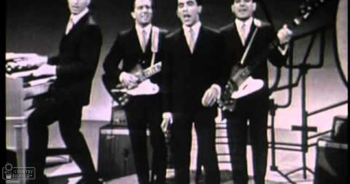 Adult males in a musical performance scene, wearing dark suits and holding microphones or playing guitars.