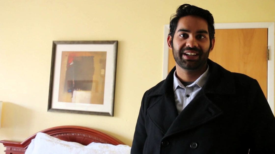 Young adult male wearing a dark overcoat with checkered shirt underneath in an indoor setting, with muted wall colors and furniture.