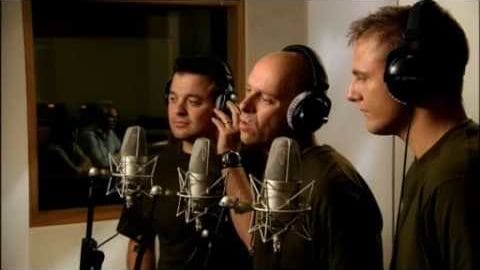 A recording studio session with three adult males, microphones and headphones in use, soundproofed wall visible, artificial lighting indoors.