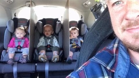 A man, likely middle-aged with a beard and mustache, wearing a plaid shirt in shades of blue, red, and white, is seated inside an airplane cabin. Three children are seated behind him; the leftmost appears to be a toddler (1-3 years old) in pink clothing, the center child seems to be an infant (<1 year), and the rightmost is another toddler wearing dark clothing.