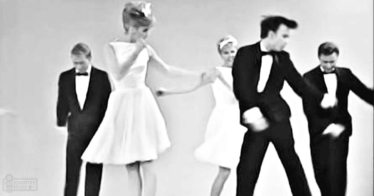 A woman in a white dress dancing, surrounded by male dancers in tuxedos against a plain white backdrop.