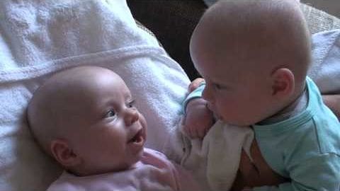 Two infants lying on a light-colored blanket or sheet in an indoor setting with neutral tones and soft lighting, likely spring or summer.