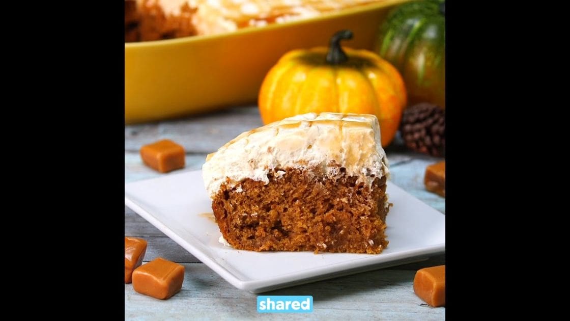 A slice of pumpkin cake with caramel drizzle on a white plate, surrounded by autumn-themed pumpkins in a kitchen setting during the daytime.