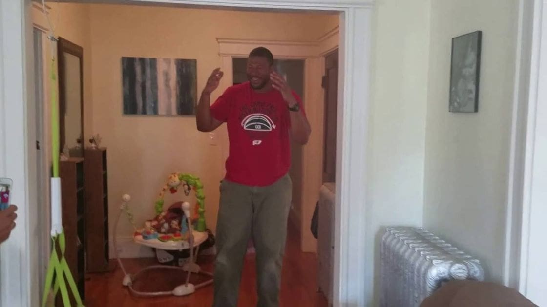 A young adult male in a residential setting wearing a red t-shirt with a graphic design, gray pants, surrounded by home decor including a baby bouncer and wall art.