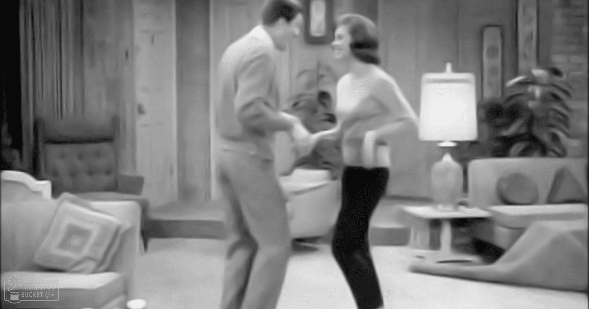 Adult male and female dancing in a mid-20th-century living room with patterned cushions and soft lighting.