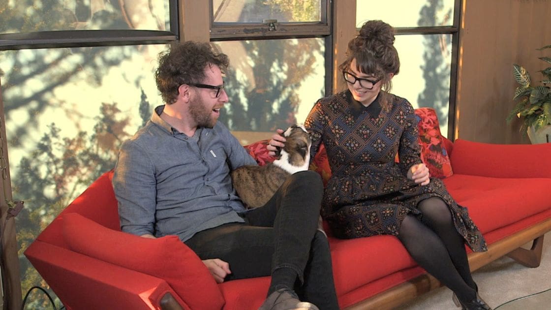 Two people sitting together on red sofas indoors; one wearing a gray button-up shirt and glasses, the other in a patterned dress with black tights and shoes; tabby cat on lap of person in shirt.