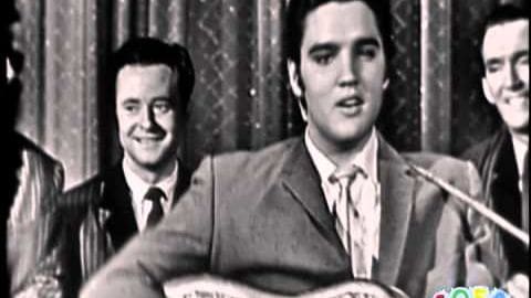 A man in his young adult years wearing a suit jacket with a tie stands indoors during what appears to be an interview or performance, accompanied by another older adult male in formal attire.