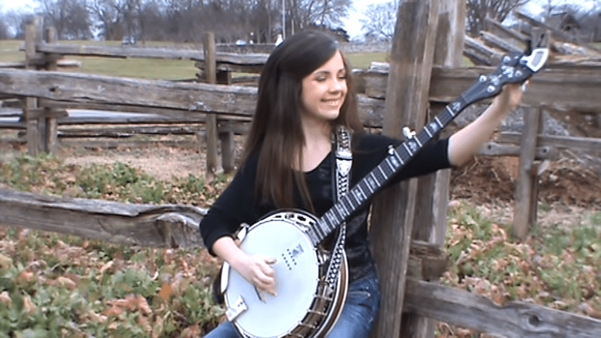 A young adult female plays a white banjo in an outdoor rural setting with wooden fences and grassy areas. The scene suggests autumn due to fallen leaves on the ground.