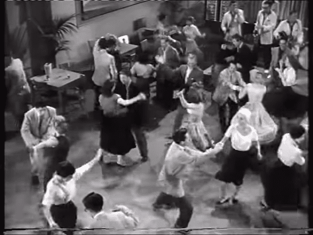 A lively dance scene with children and adults gathered indoors, possibly at a social gathering or party; tables with objects suggesting drinks or food items are visible.