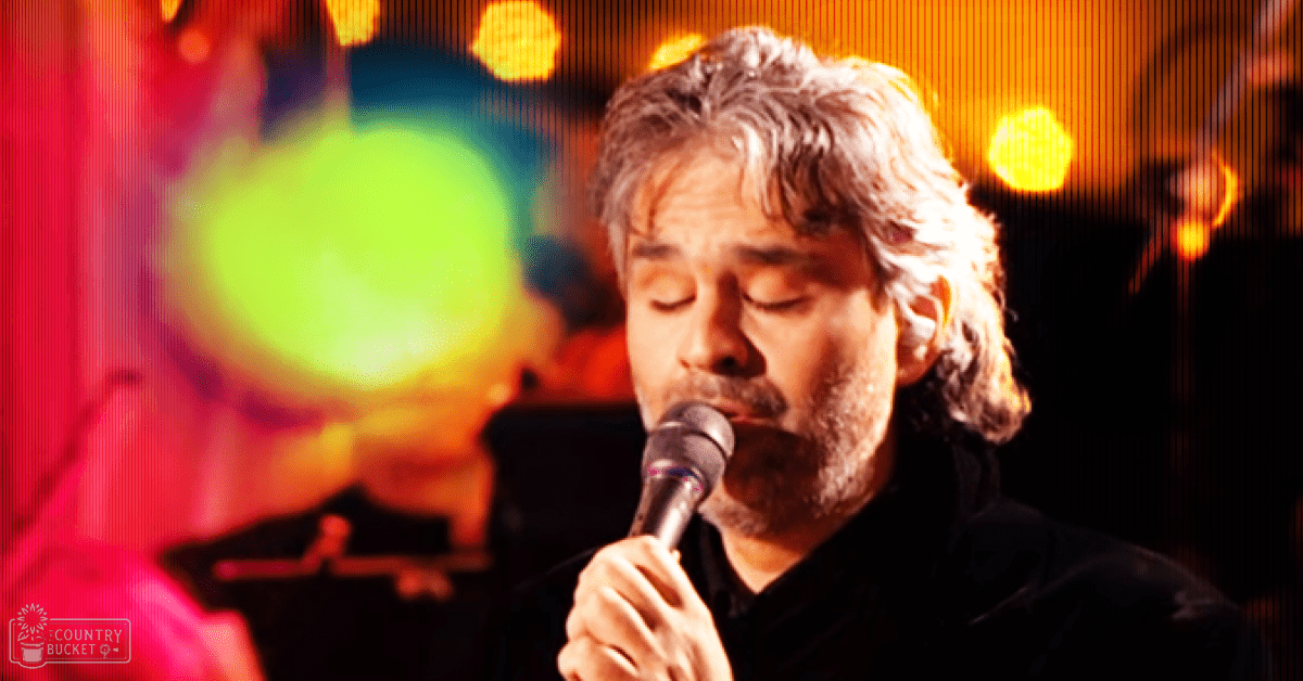 A middle-aged male singer performing into a microphone at what appears to be a concert setting with stage lighting and musical instruments in the background.