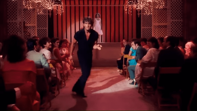Adult male dancing on stage at an indoor event space, surrounded by seated adults in various attire during what appears to be a formal gathering.
