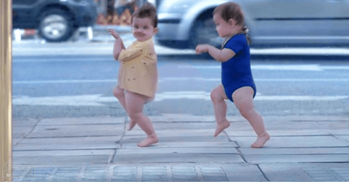 Two toddlers wearing colorful outfits walk along a sidewalk near an urban street with vehicles visible in the background. The scene is bright and sunny, suggesting spring or summer.