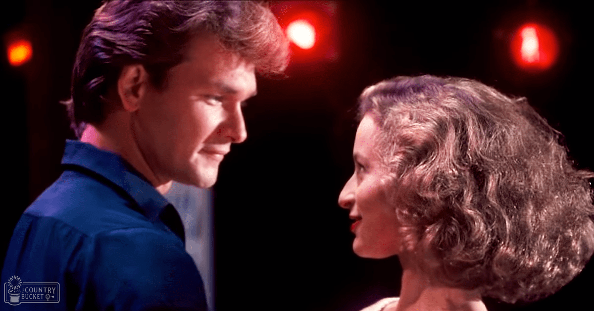 A couple in an intimate dance scene, possibly from a film, set against a dark stage backdrop with red circular lights resembling emergency exit signs or stage lighting.