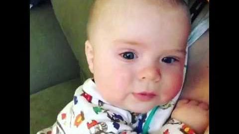 An infant wearing a white, hooded onesie with bright, playful cartoon patterns, seated indoors near green upholstered furniture. Natural light suggests daytime in an everyday indoor setting.