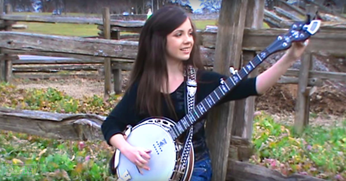 A young adult female plays a banjo in an autumnal outdoor setting with wooden fencing, fallen leaves, and a stone structure.