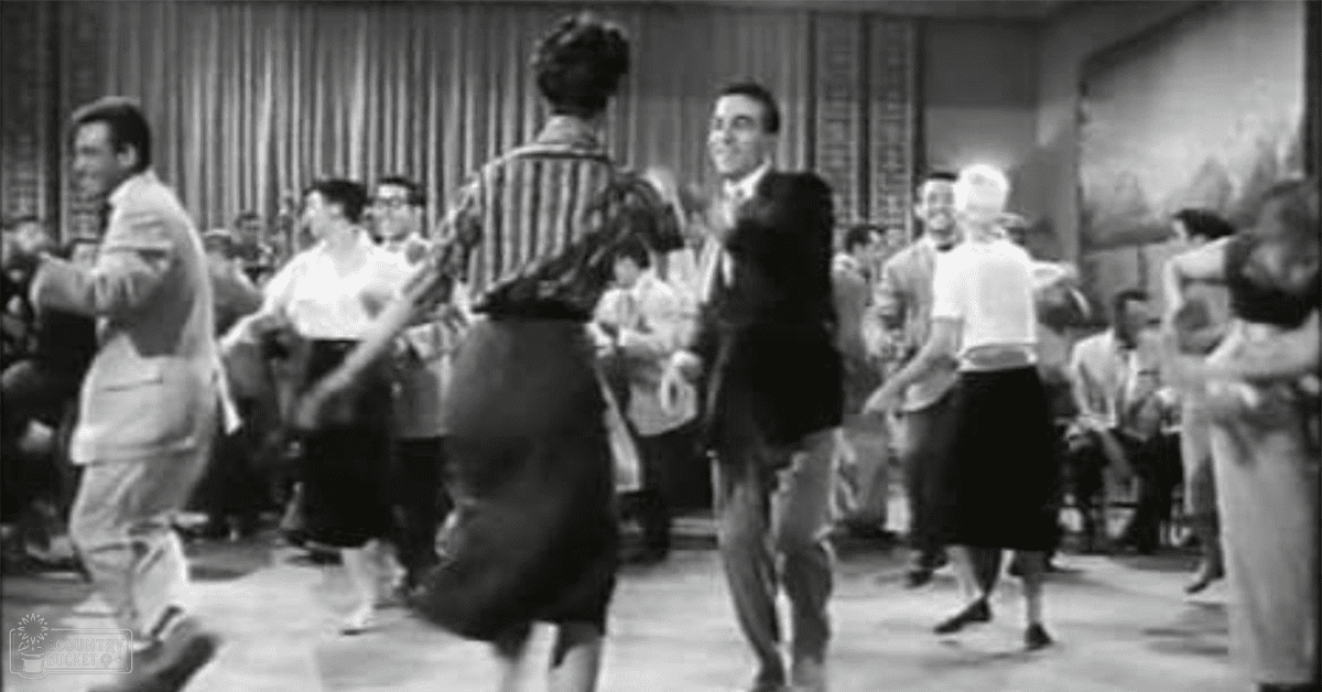 A dance scene with young adults in a mid-20th-century setting with wooden paneling and curtains.