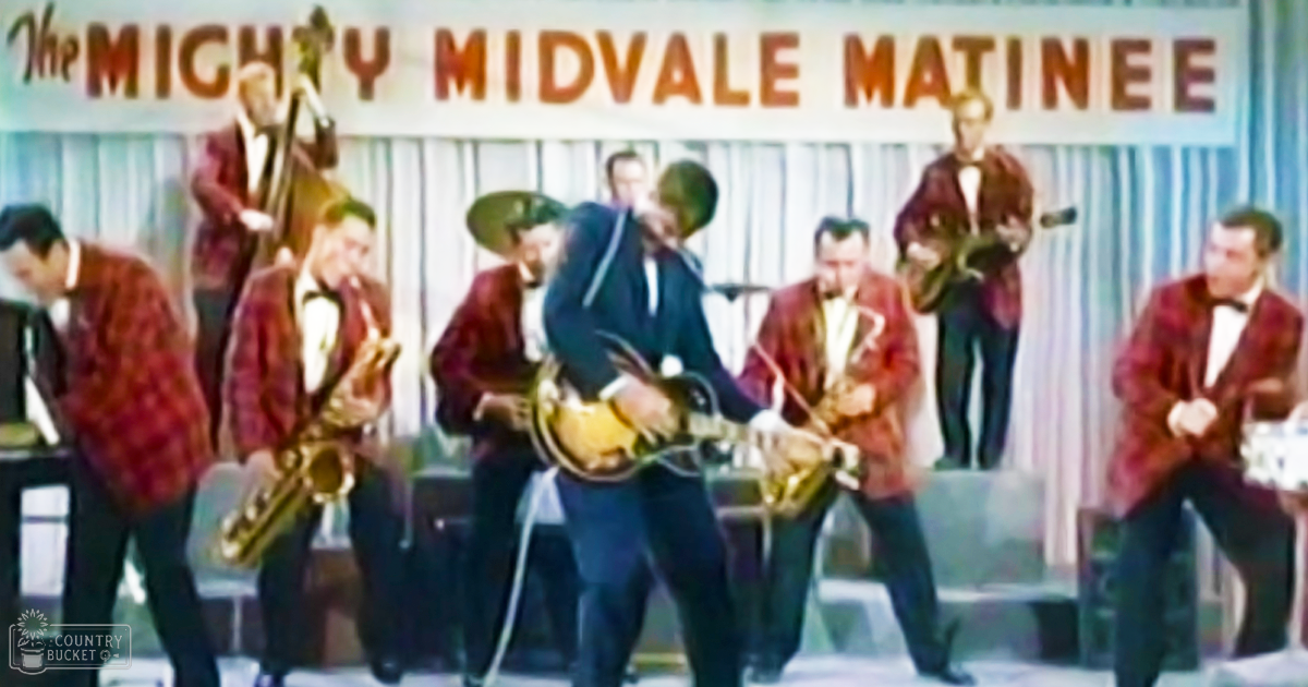 A musical performance at 'The Mighty Midvale Matinee' with adult male performers wearing red plaid blazers and dark suits, set on an indoor stage with musical instruments.