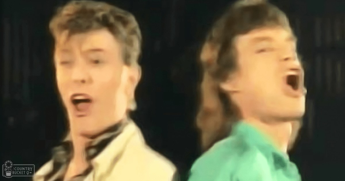 Two adult males in a dimly lit performance setting, one wearing a light-colored button-up shirt and the other in a teal t-shirt with long sleeves and short hair tied back.