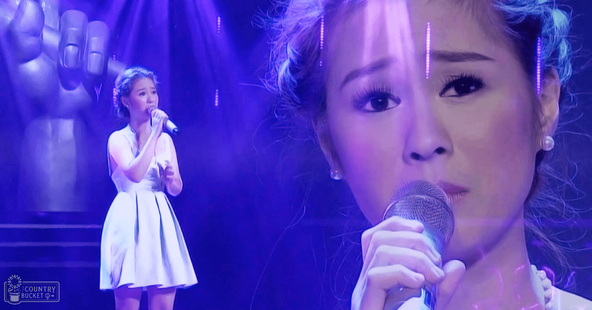 Young adult female singer in a white sleeveless dress performing on an indoor concert stage with stage lighting and a large hand sculpture behind her.