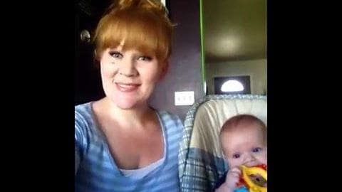 A middle-aged woman with red hair holds an infant indoors; she wears a blue and white striped top, while the infant is dressed in light-colored attire. The setting includes indoor furniture with a plaid pattern.