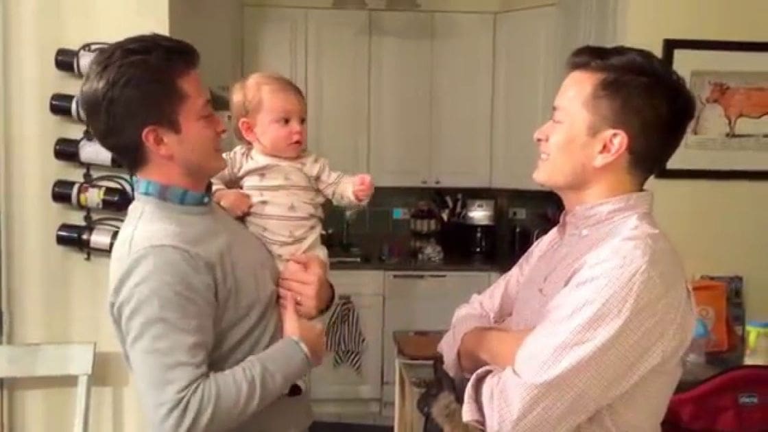 A baby being held by two adults in a modern kitchen with white cabinets and a wine rack, natural light streaming through an unseen window.