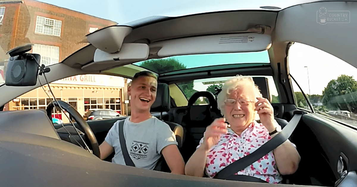Young adult male driver wearing a light gray t-shirt with an Aztec-inspired pattern, accompanied by an older female passenger in a white blouse with pink floral patterns inside a car parked near 'China Grill' restaurant during the daytime.