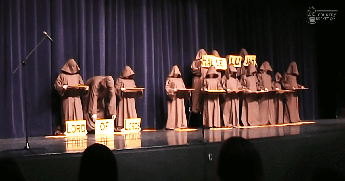 A group performance on a dark-curtained indoor stage with young male adults wearing loose, brownish-tan hooded robes during evening lighting.