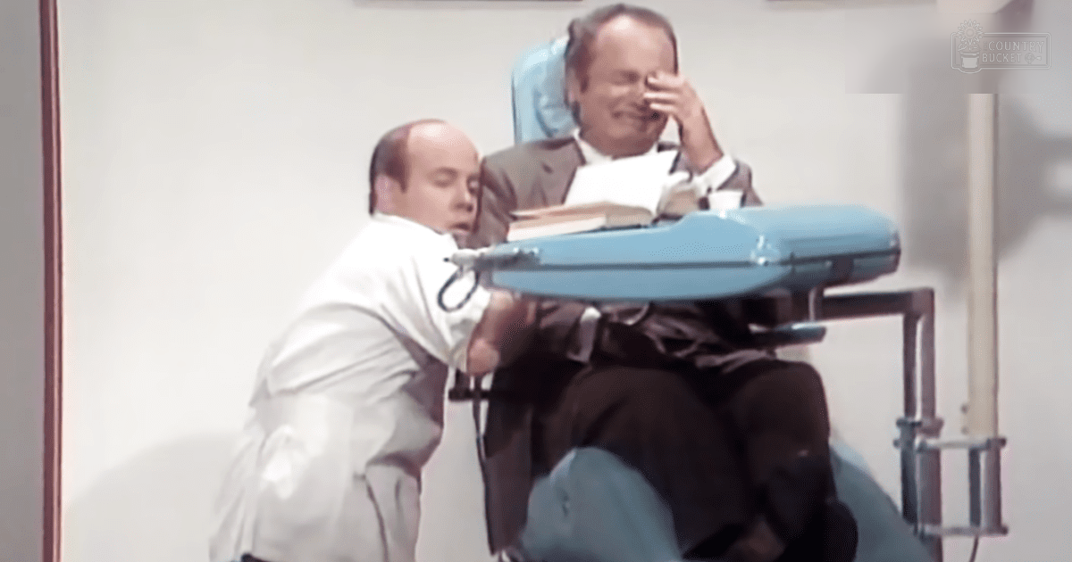 An older adult male in a dark suit sits at an examination table, while a middle-aged to older adult male doctor wearing a white lab coat stands beside him in what appears to be a clinical office setting with artificial lighting.