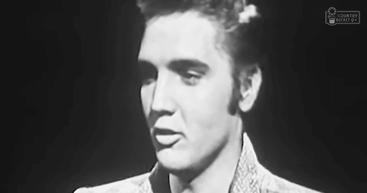 A young adult male in a relaxed, casual collared shirt with a subtle pattern from what appears to be the mid-20th century stands against a dark background suggesting stage or performance lighting.