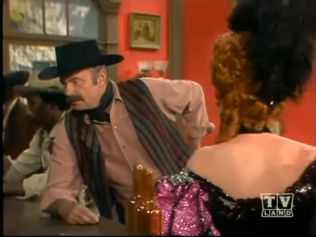 A middle-aged man wearing a wide-brimmed black cowboy hat and light brown rolled-up-sleeve shirt, seated in what appears to be an indoor bar or saloon with a red wall and framed pictures.