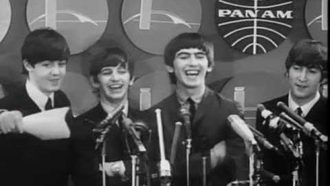 A group of young adult males at a press conference or public speaking event, dressed in dark suits from the 1960s era, with microphones and 'PAN AM' backdrop visible.