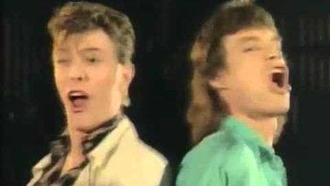 Two young adult males performing on stage, one wearing a plaid shirt and the other a light green t-shirt, with dark curtains as background.
