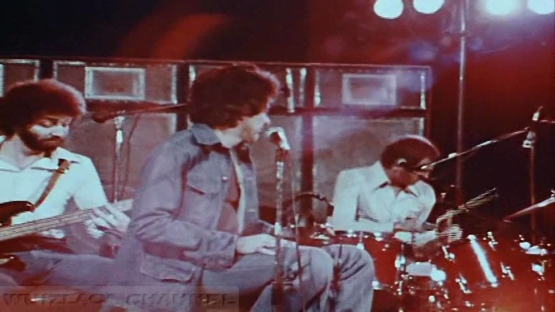 Musical performance with adult males playing electric guitar and drums in a stage setting with bright artificial lighting.