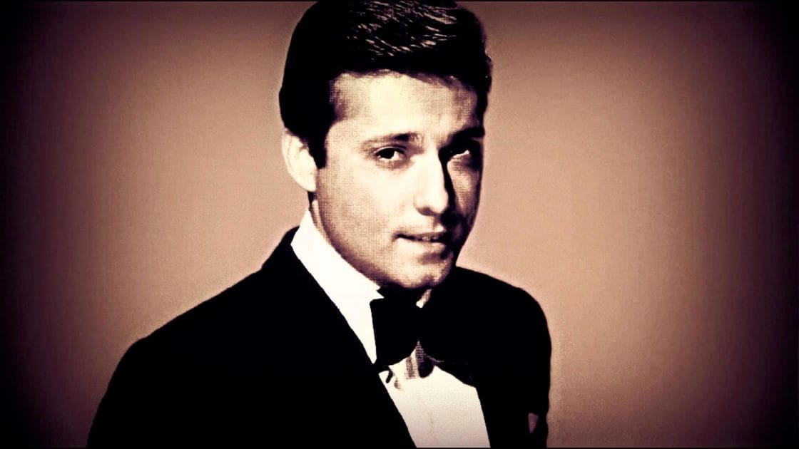A young adult male wearing a formal suit with a bow tie stands against plain indoor walls, likely during an event from the mid-20th century.